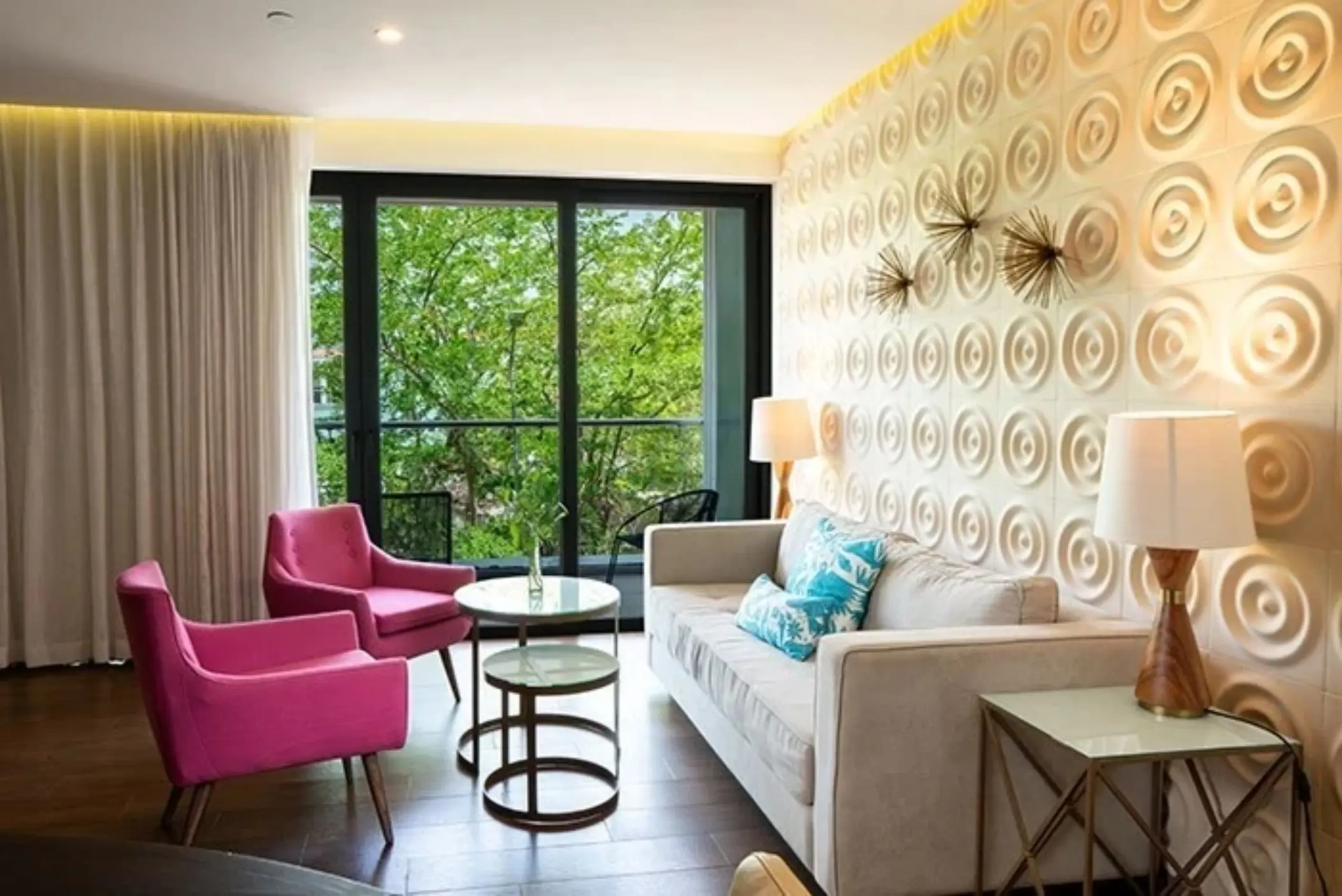 Bright living area with cream sofa, pink chairs, glass tables, and balcony views surrounded by lush greenery.