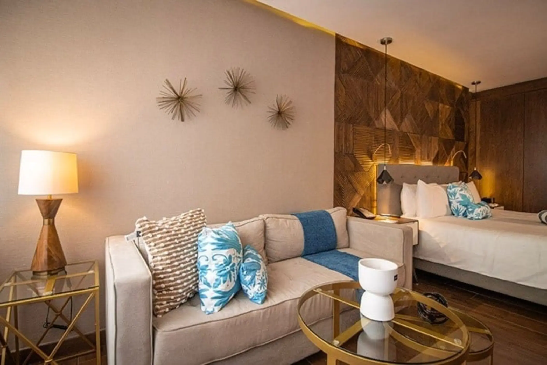 Cozy hotel room with neutral sofa, blue pillows, and a king bed against a wooden geometric wall with warm lighting.