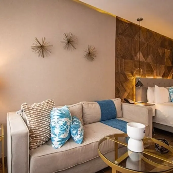 Cozy hotel room with neutral sofa, blue pillows, and a king bed against a wooden geometric wall with warm lighting.