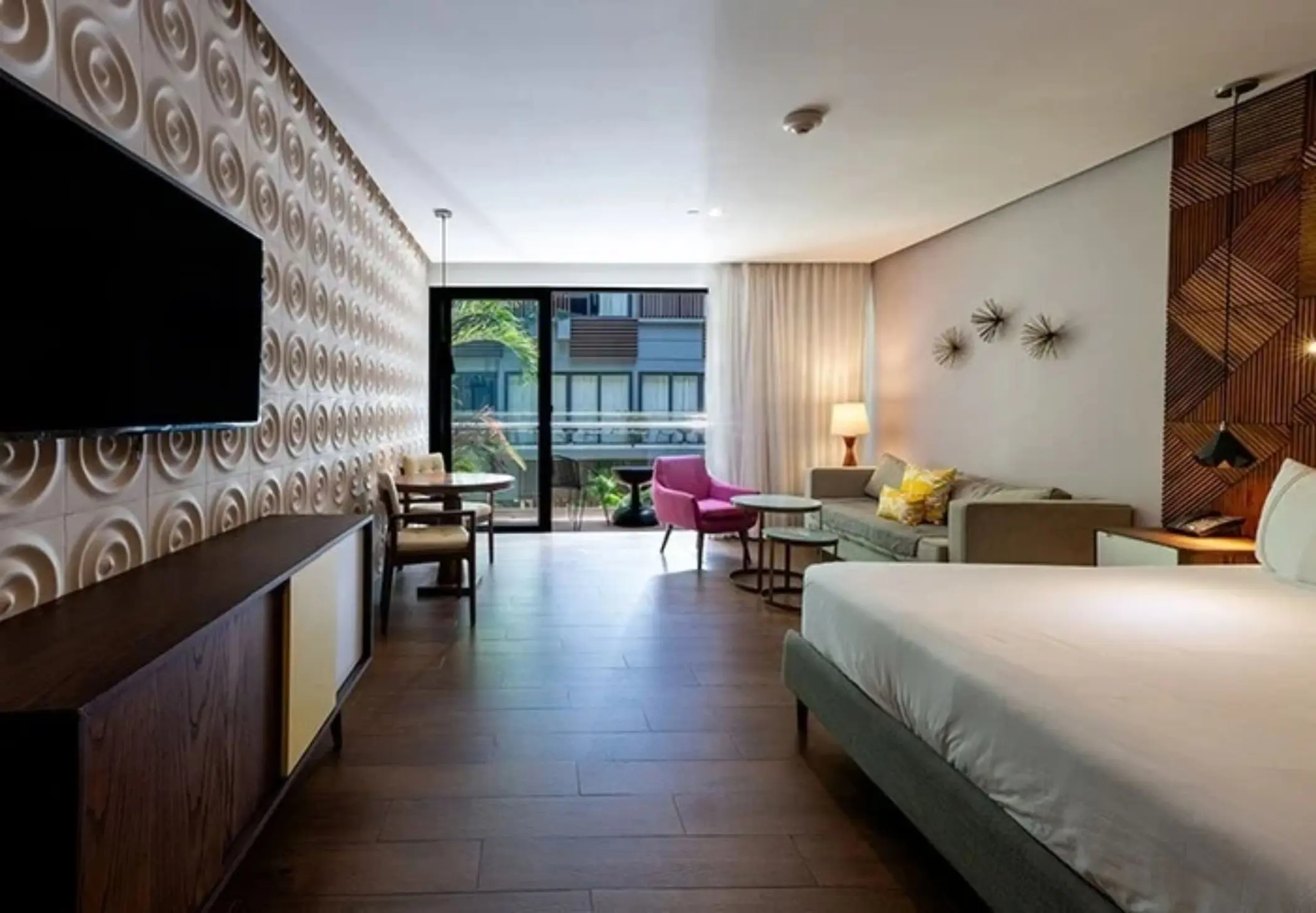 Open-plan suite with wood floors, a king bed, and modern furnishings, plus balcony view of courtyard greenery.