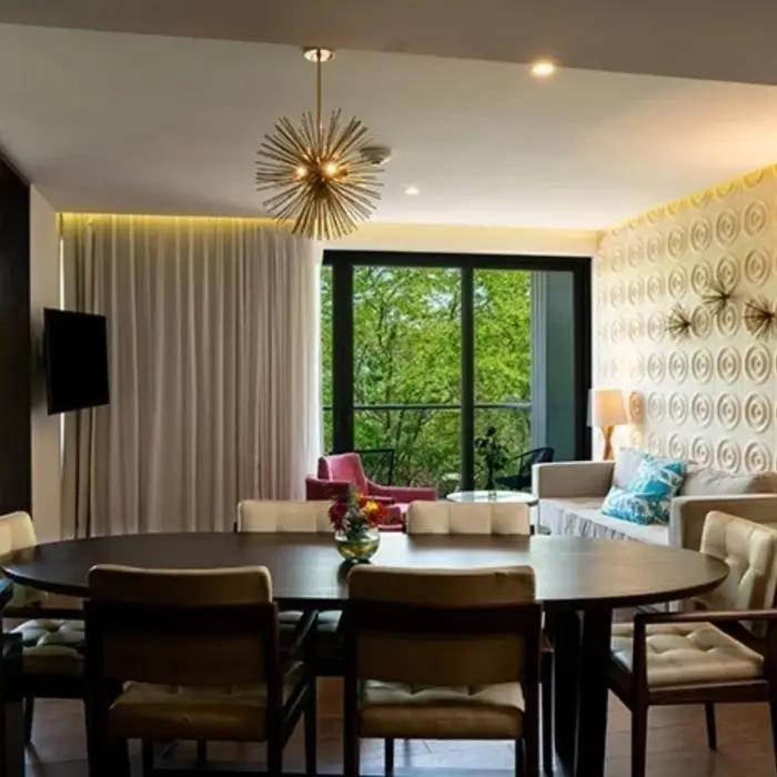 Large dining area with modern light fixture, plush chairs, and balcony view of lush greenery through glass doors.