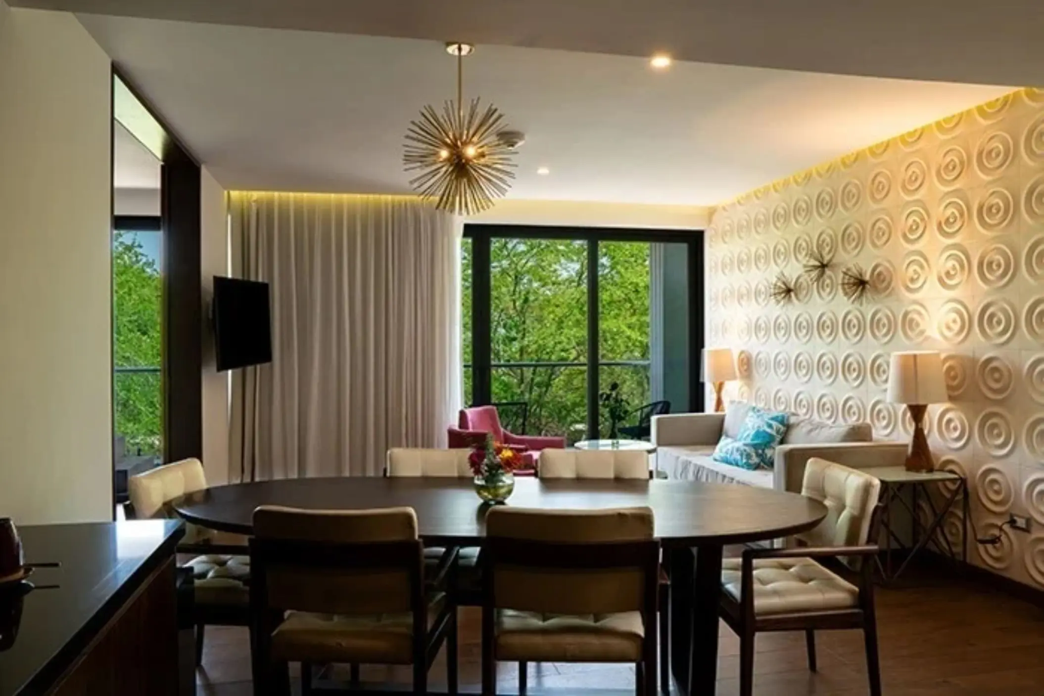 Large dining area with modern light fixture, plush chairs, and balcony view of lush greenery through glass doors.