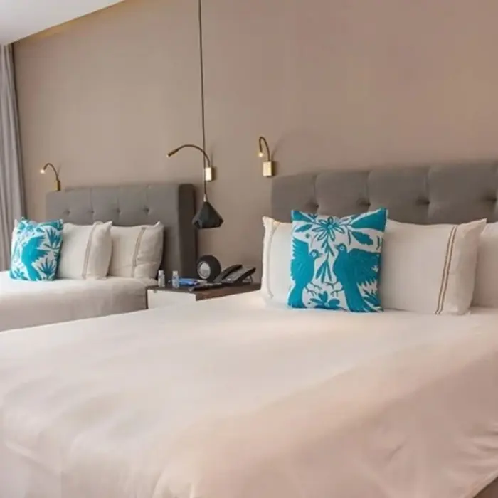 Bright room with two double beds, teal embroidered pillows, and natural light filtering through soft white curtains.