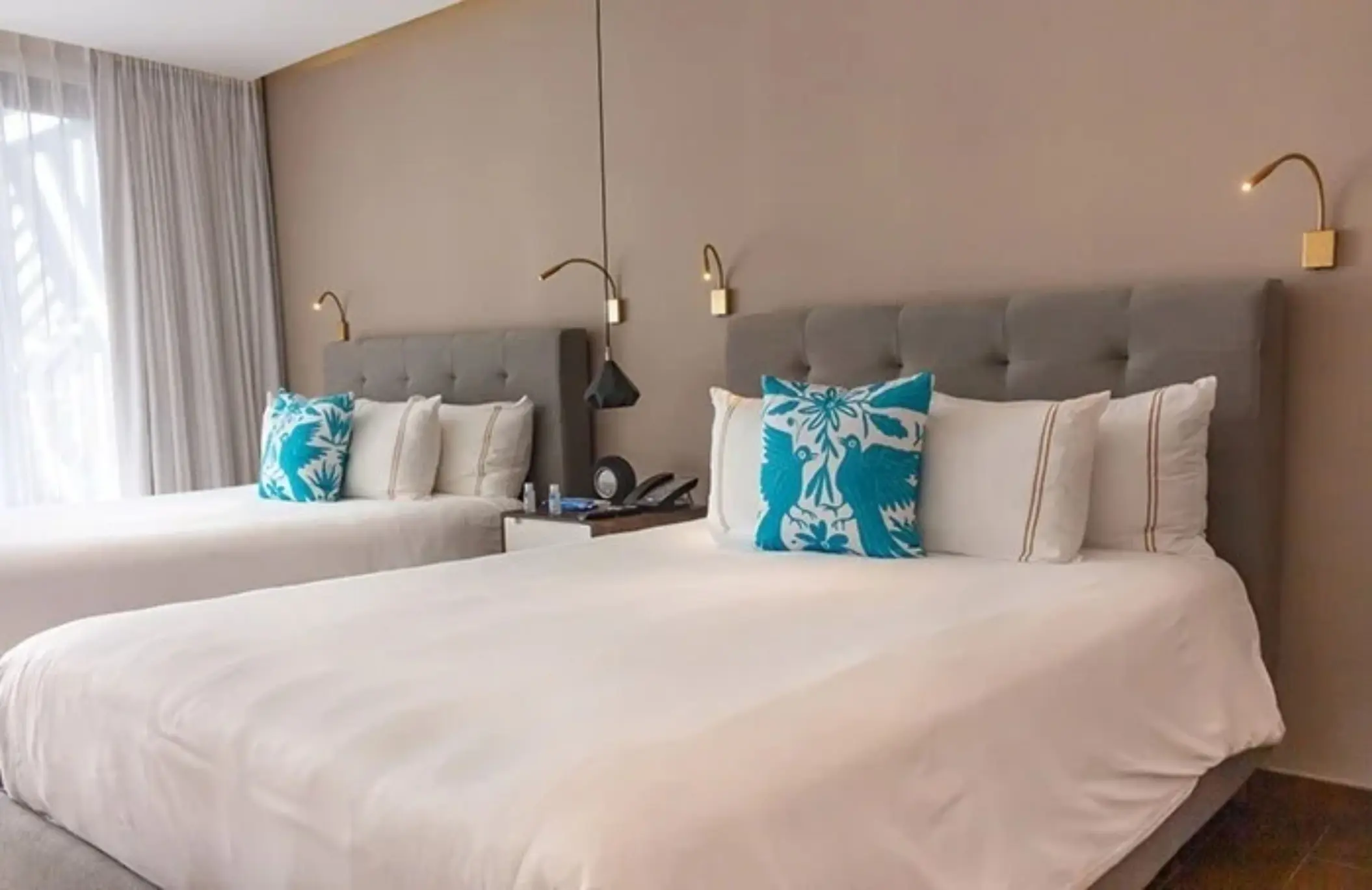 Bright room with two double beds, teal embroidered pillows, and natural light filtering through soft white curtains.