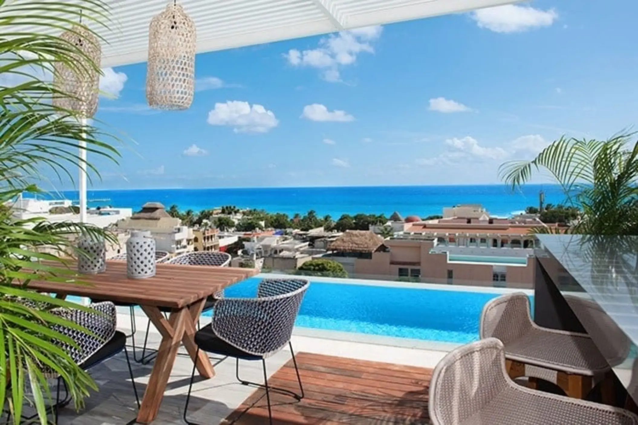 Rooftop terrace with plunge pool, ocean view, wooden table, and stylish woven chairs under hanging lanterns.