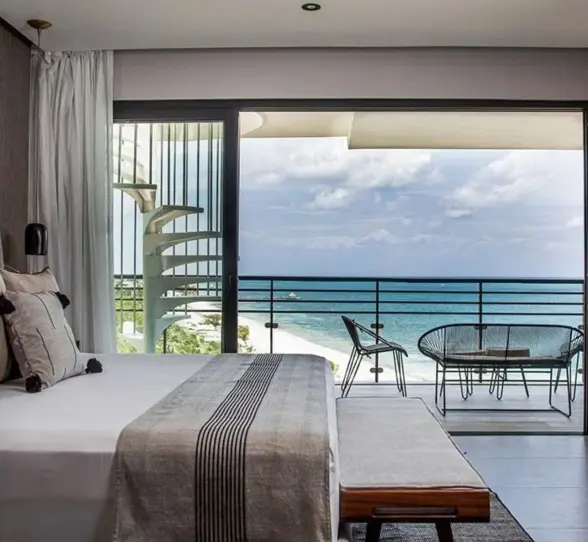Oceanview bedroom with king bed, private balcony, and spiral staircase overlooking a white sand beach and turquoise sea.
