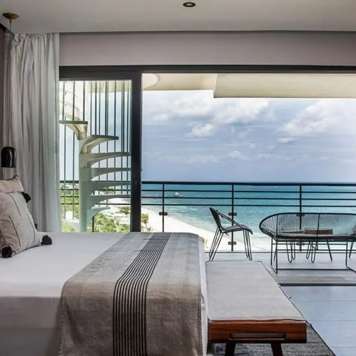 Oceanview bedroom with king bed, private balcony, and spiral staircase overlooking a white sand beach and turquoise sea.