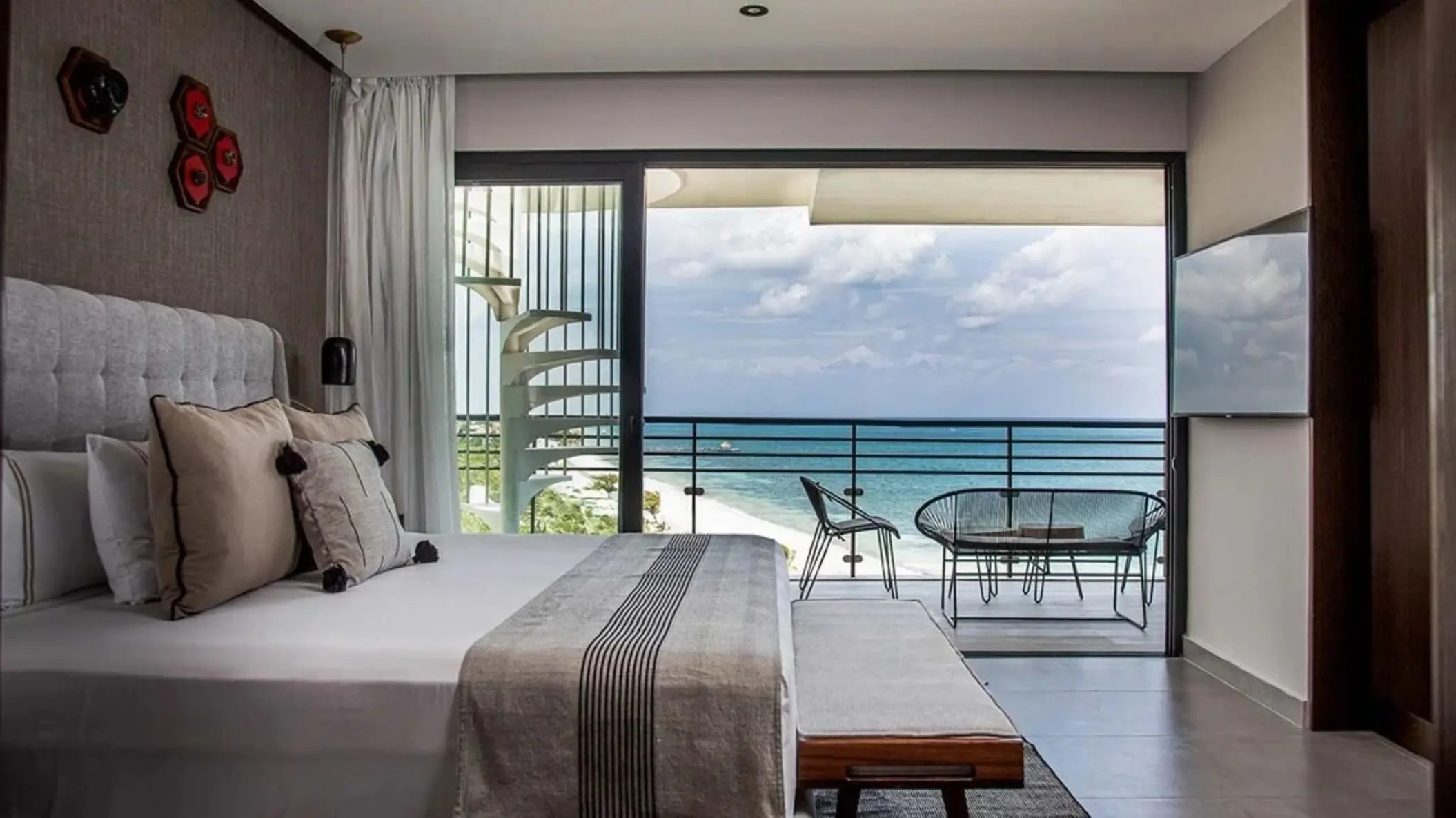 Oceanview bedroom with king bed, private balcony, and spiral staircase overlooking a white sand beach and turquoise sea.