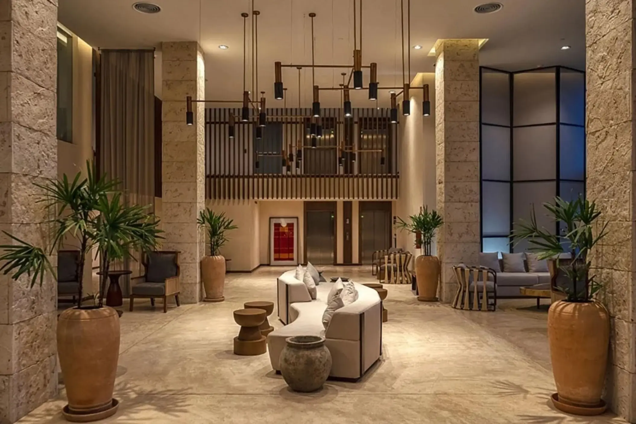 Spacious hotel lobby with modern lighting, natural stone pillars, large potted plants, and cozy seating areas.