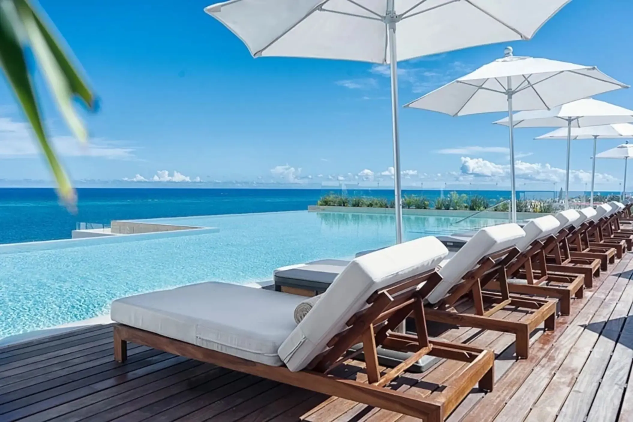 Infinity pool with white umbrellas and wooden loungers facing the bright blue Caribbean Sea under a clear sky.