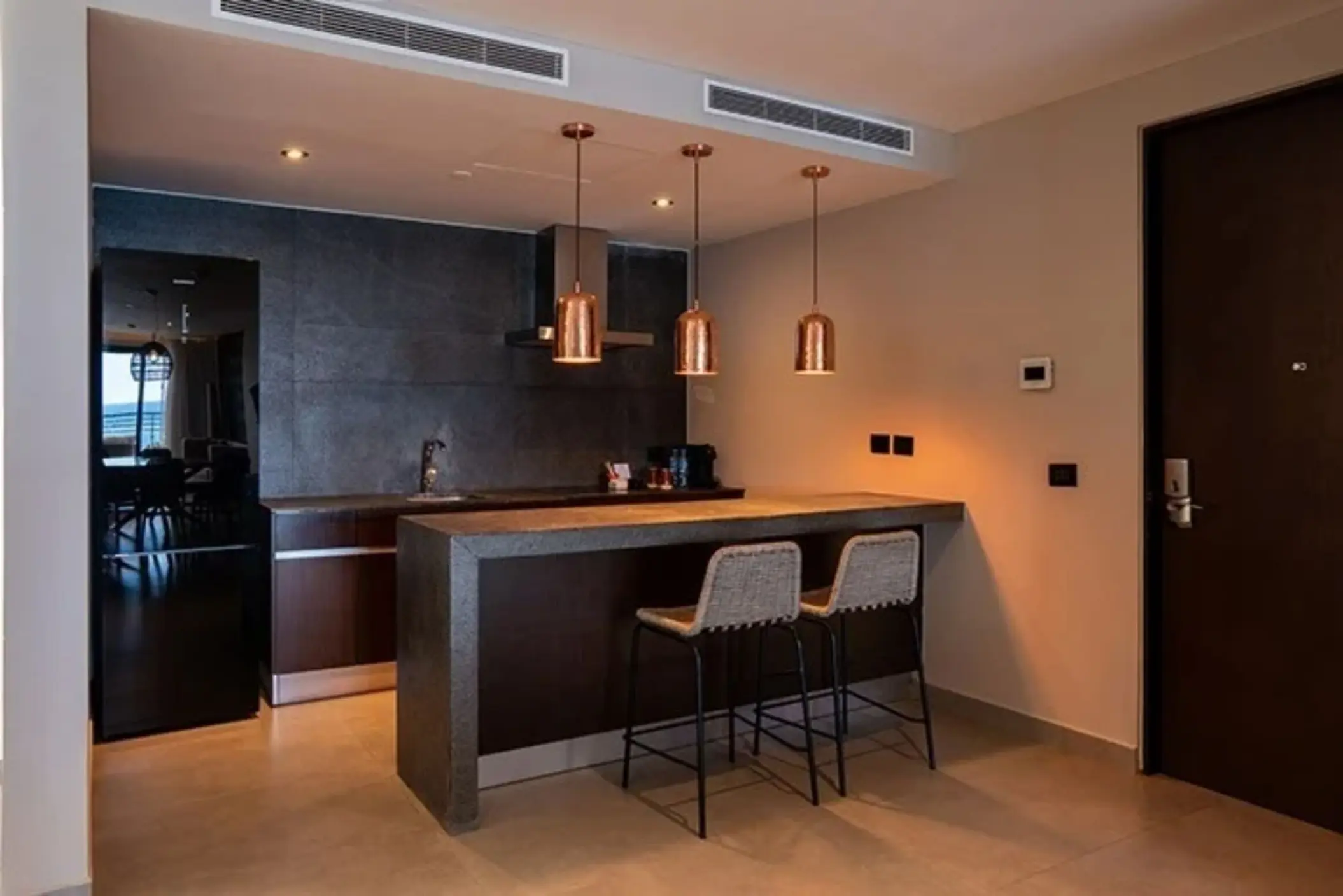 Stylish kitchenette with dark stone countertops, copper pendant lights, and two woven barstools.