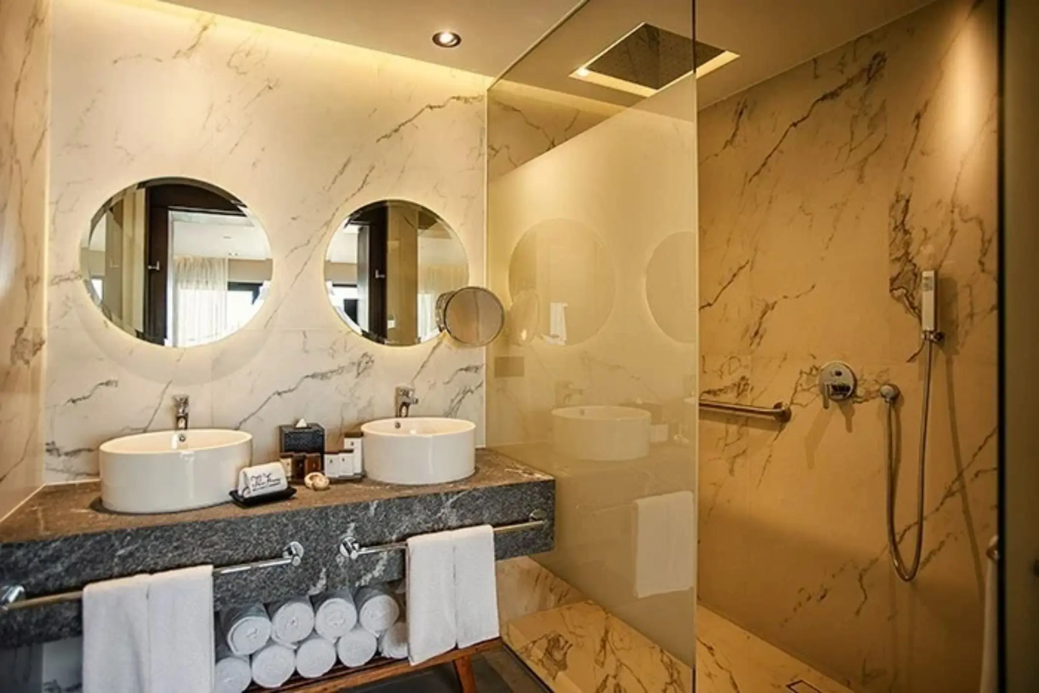 Marble bathroom with dual sinks, round mirrors, walk-in rain shower, and elegant lighting.