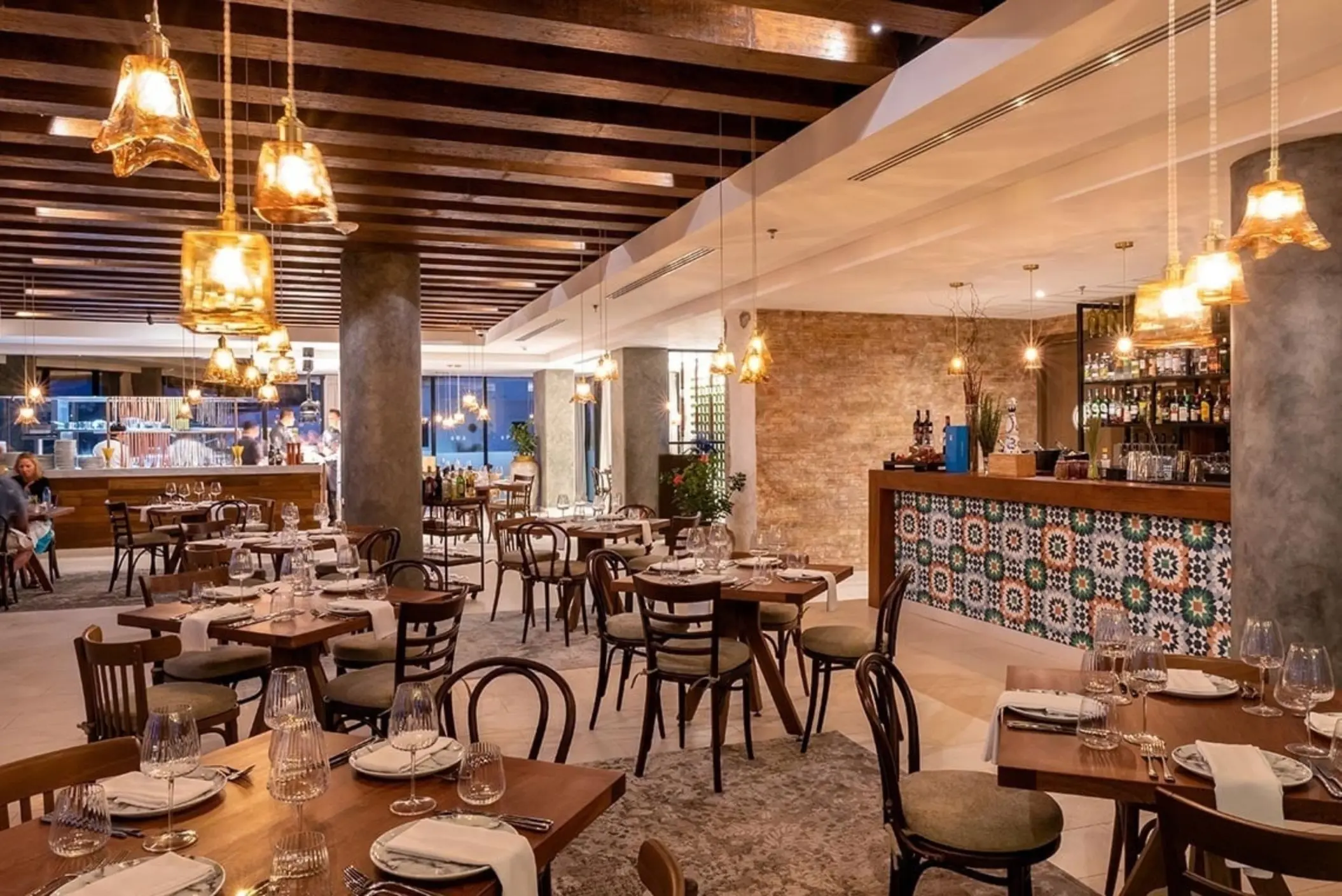 Elegant restaurant with warm lighting, wood beam ceiling, and colorful tiled bar surrounded by rustic decor.