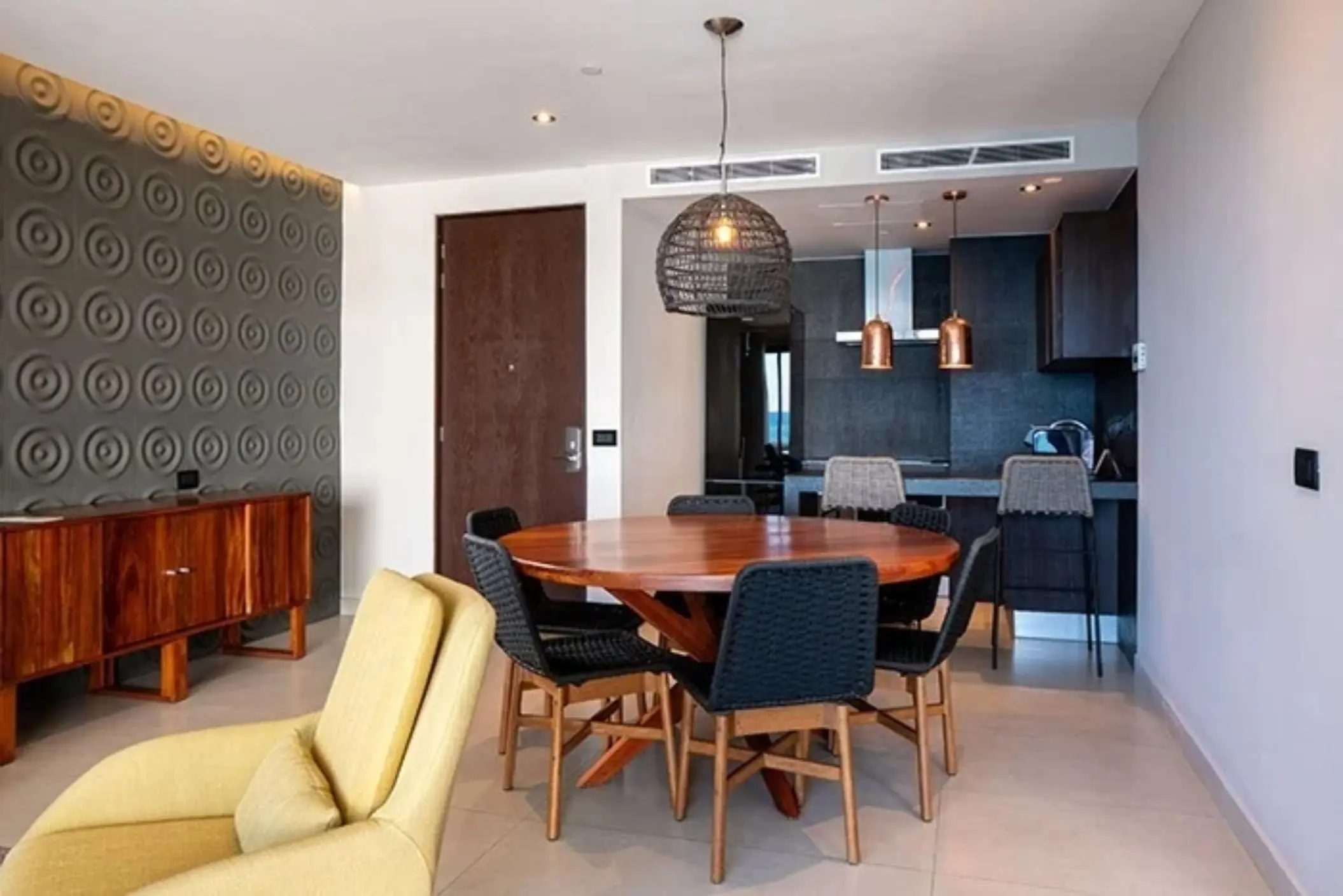 Cozy dining area with round table, woven pendant light, and kitchenette featuring copper accents and barstools.