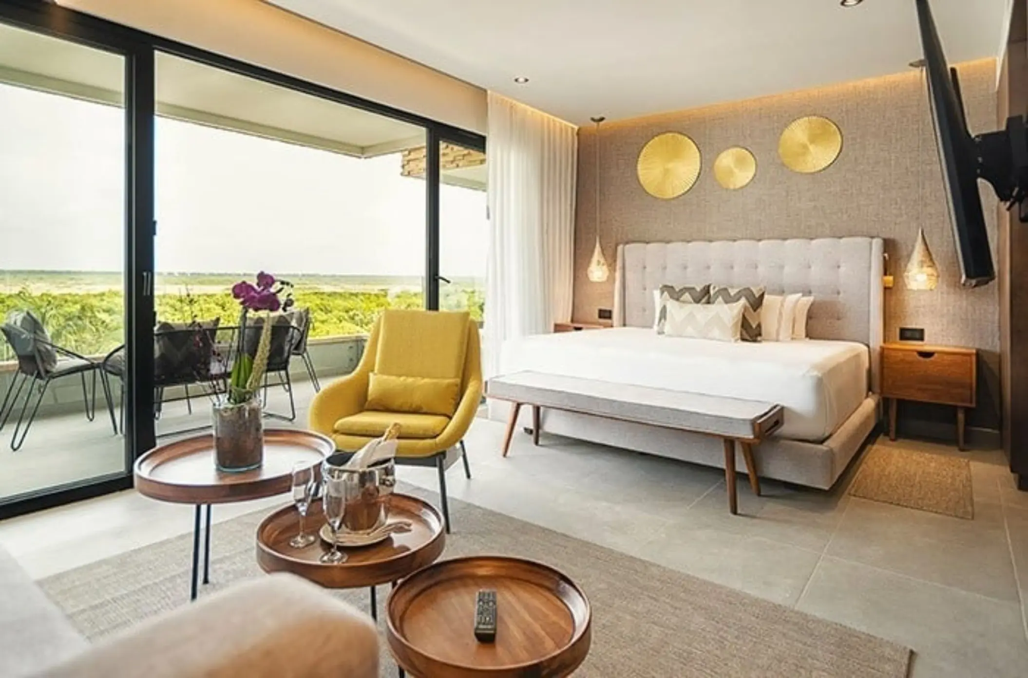 Stylish bedroom with king bed, yellow chair, balcony seating, and golden wall decor with a view of greenery.
