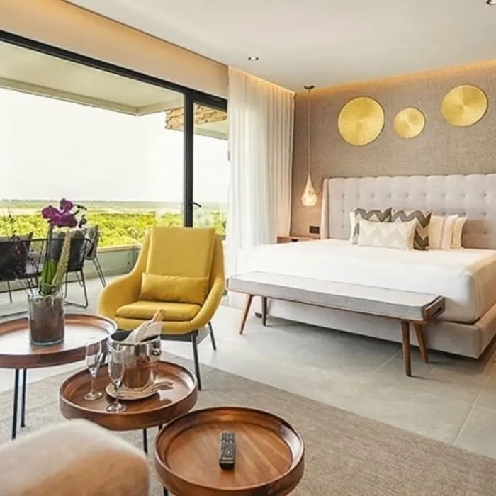 Stylish bedroom with king bed, yellow chair, balcony seating, and golden wall decor with a view of greenery.