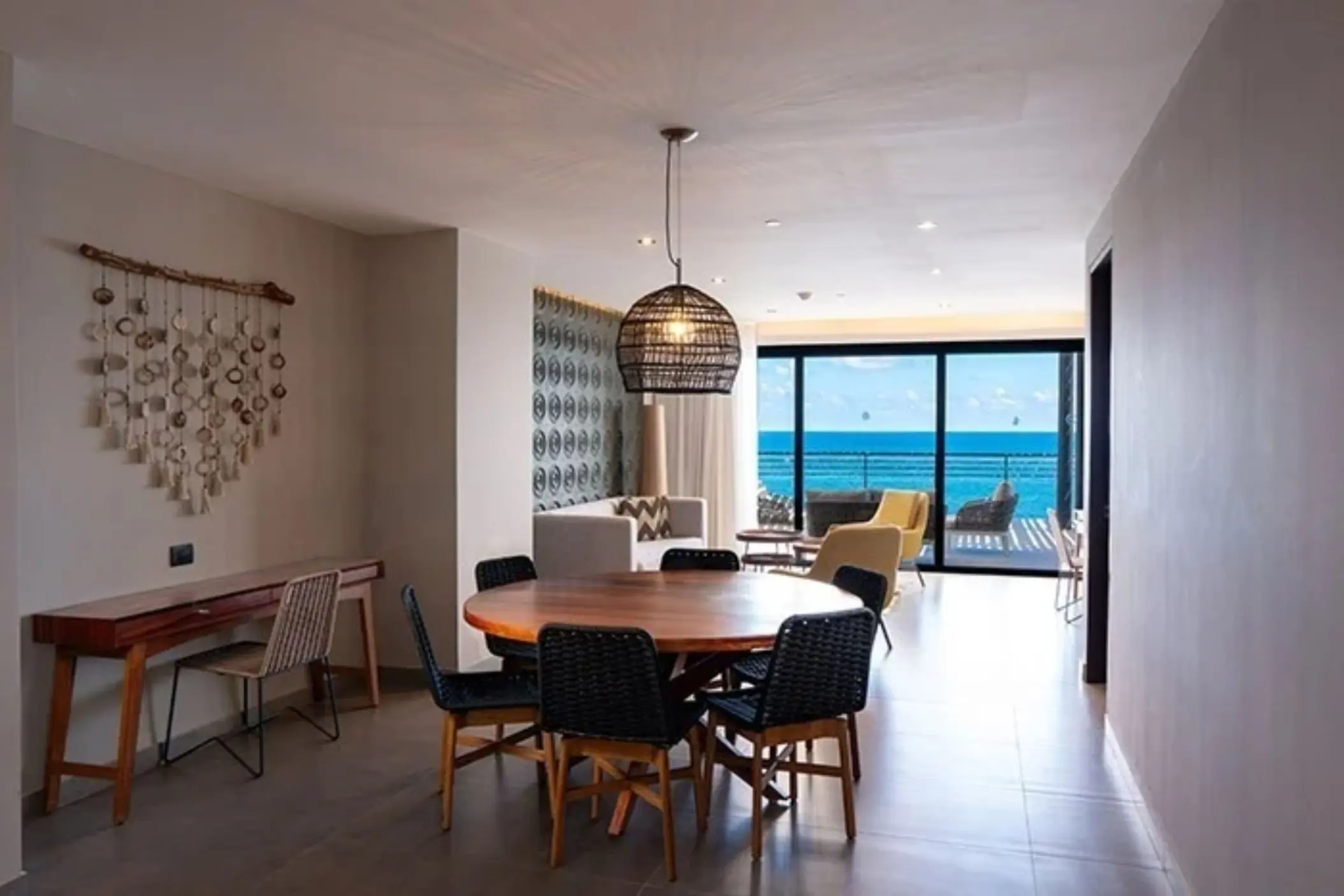 Dining area with woven pendant light and ocean view balcony in the distance through glass doors.