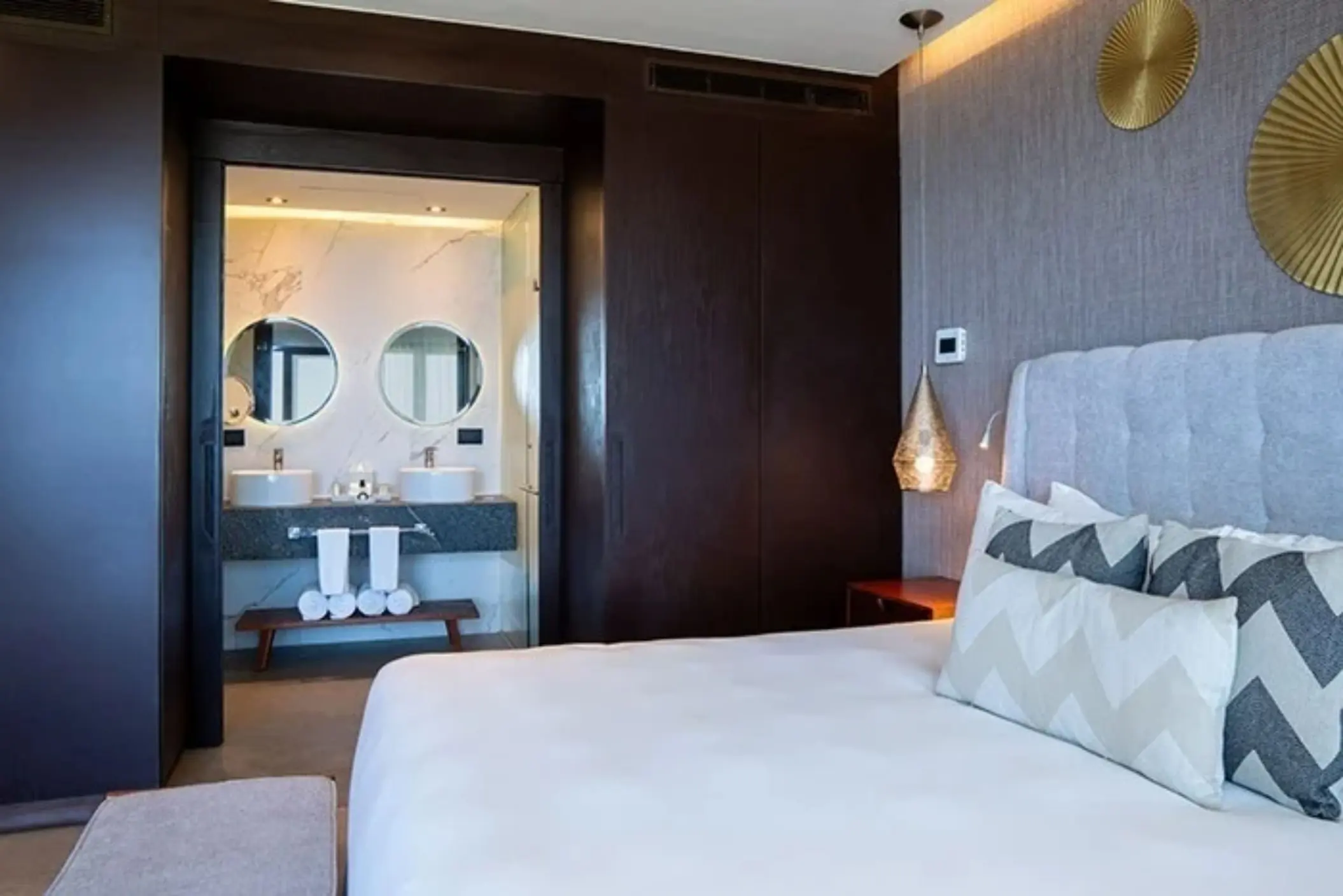 Modern bedroom with king bed, dark wood accents, and open view into a double vanity bathroom.