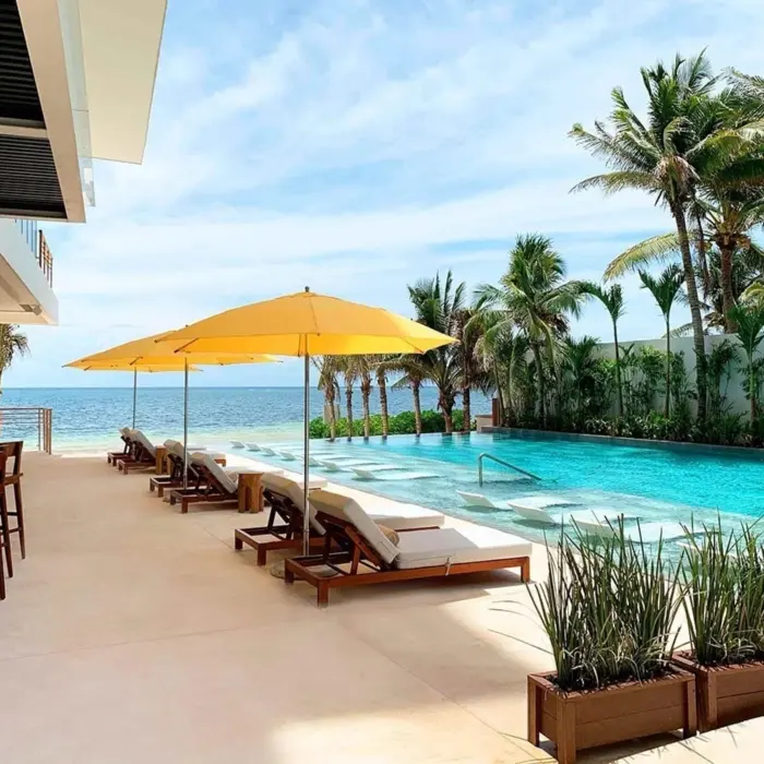 Infinity pool with yellow umbrellas and sun loungers facing the ocean, surrounded by tropical palms and open bar area.