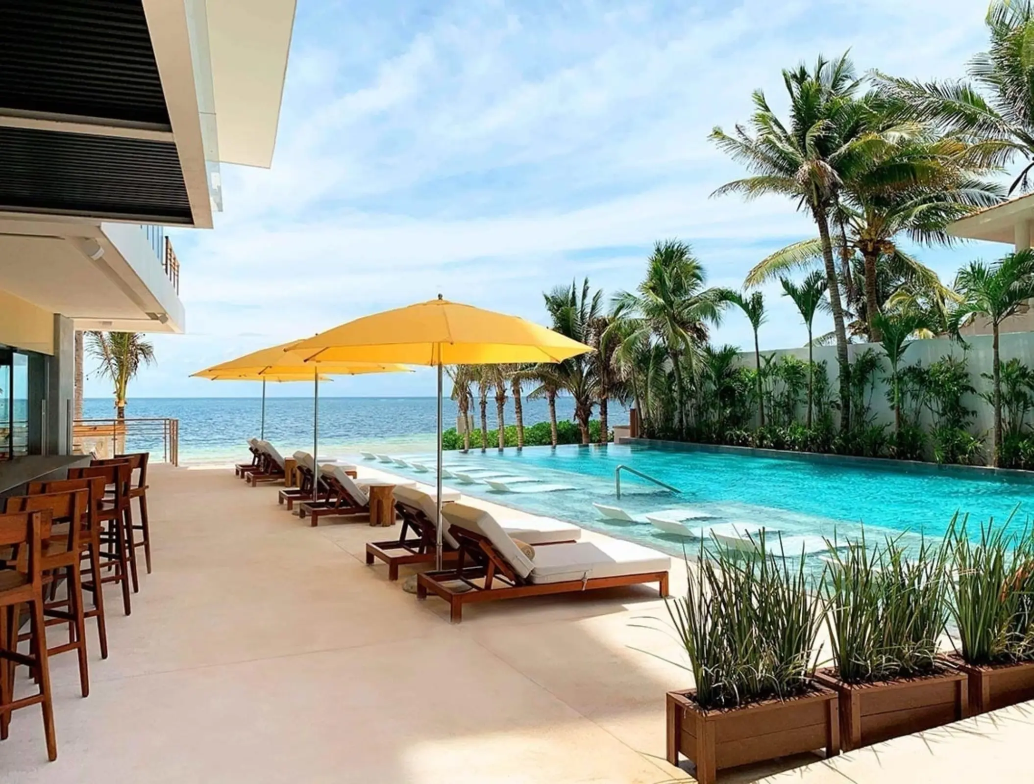 Infinity pool with yellow umbrellas and sun loungers facing the ocean, surrounded by tropical palms and open bar area.