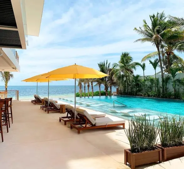 Infinity pool with yellow umbrellas and sun loungers facing the ocean, surrounded by tropical palms and open bar area.