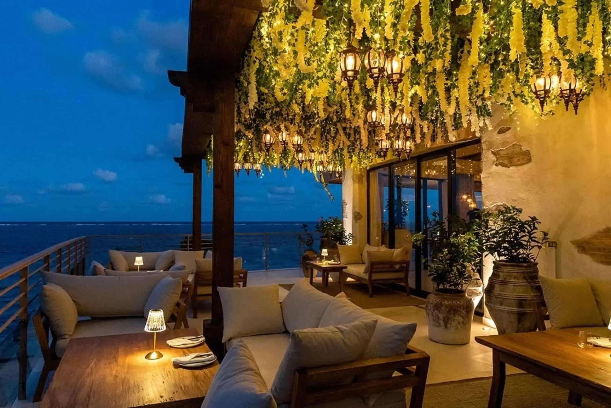 Romantic oceanview terrace with cozy seating, lantern lighting, and hanging greenery glowing under the evening sky.