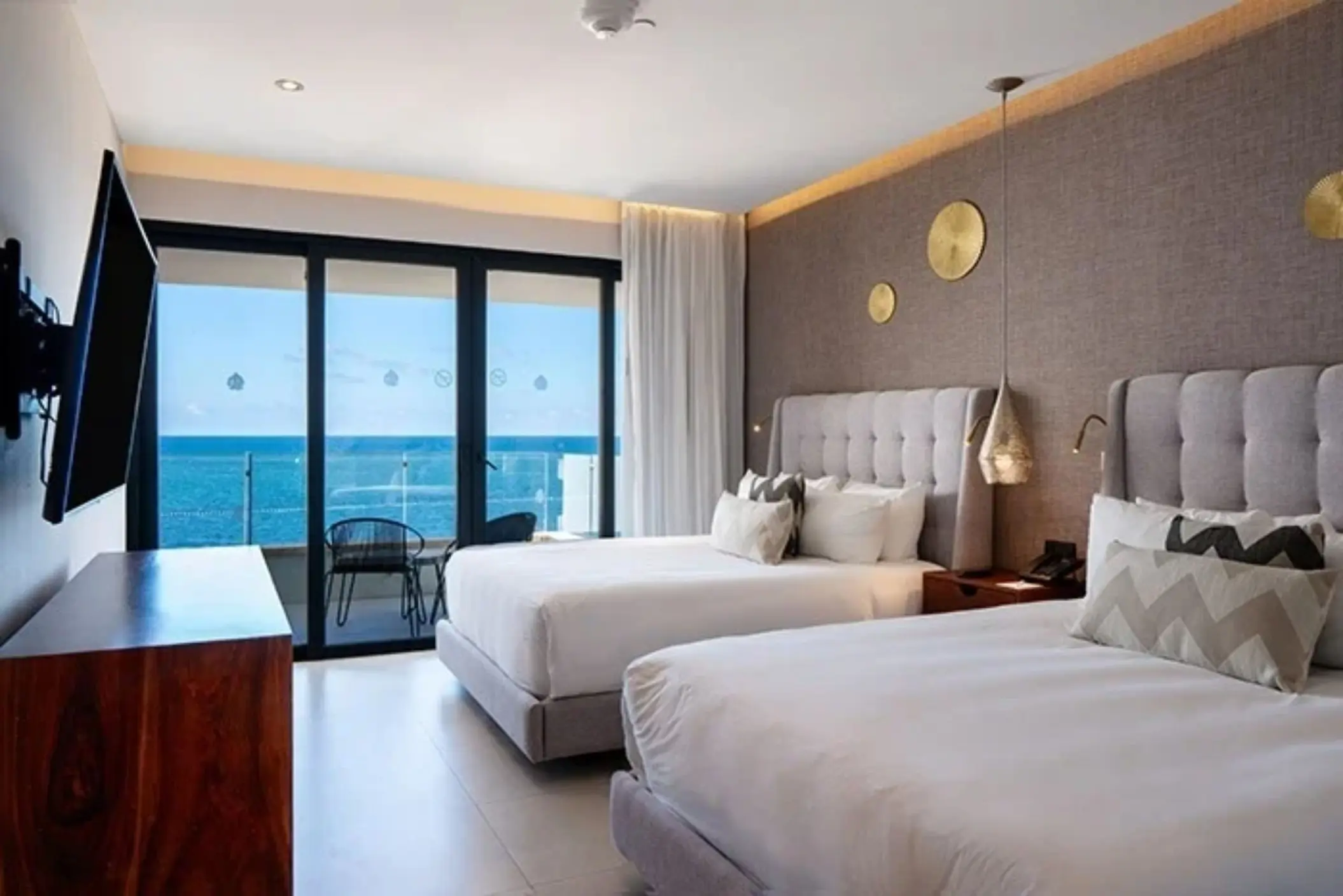 Contemporary room with two queen beds and private balcony overlooking the ocean through floor-to-ceiling glass doors.