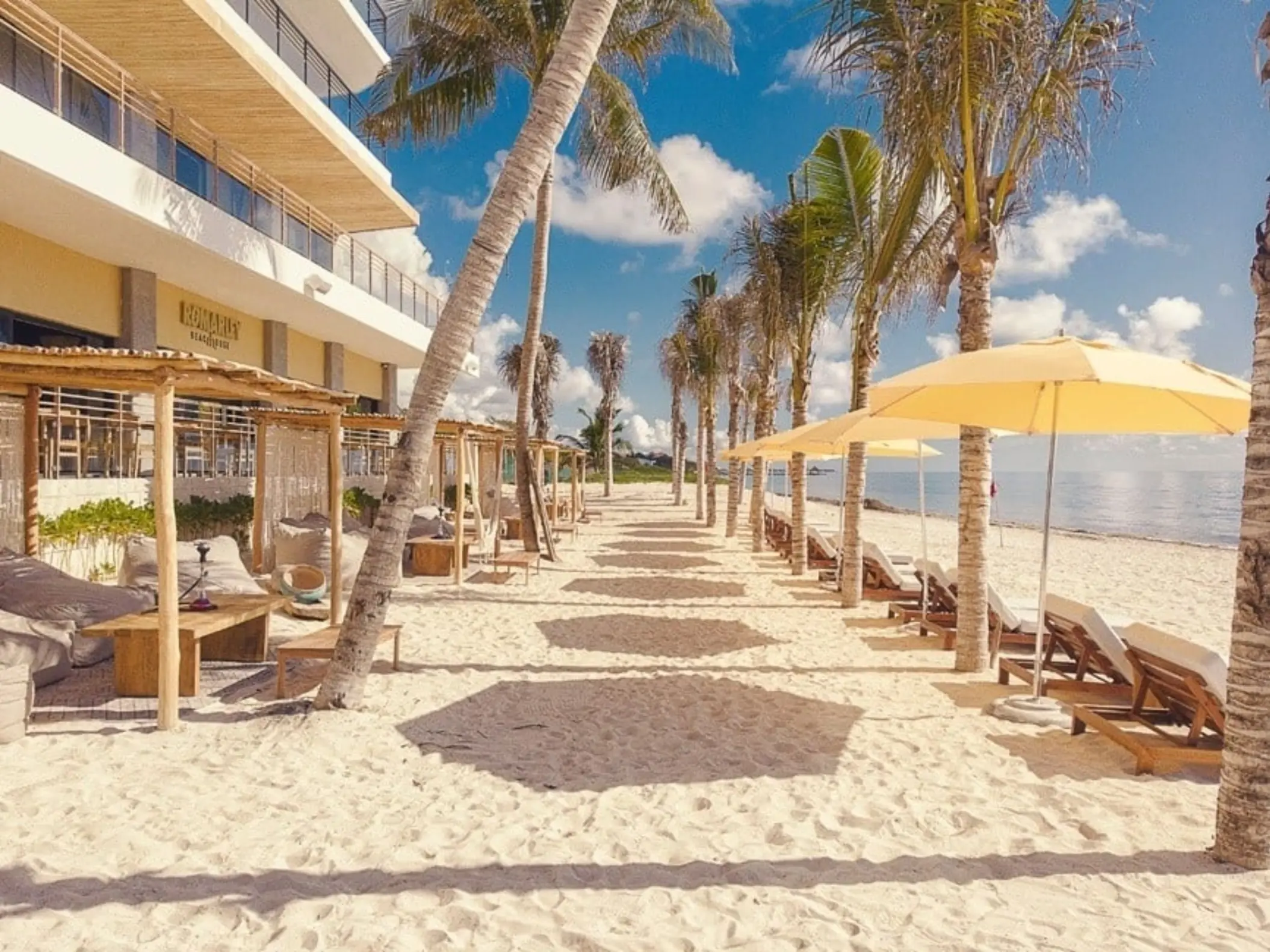 Sandy beachfront with loungers, cabanas, and palm trees outside a resort lined with umbrellas and ocean views.