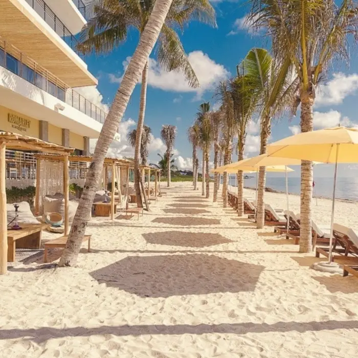 Sandy beachfront with loungers, cabanas, and palm trees outside a resort lined with umbrellas and ocean views.