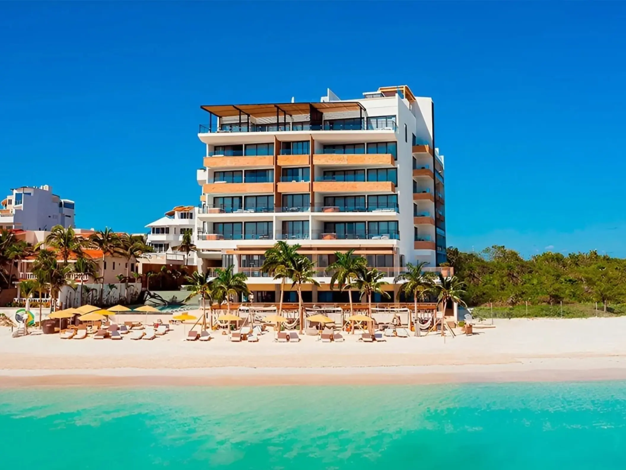 Beachfront resort with modern design, surrounded by palm trees and loungers, directly on a white sand shoreline.