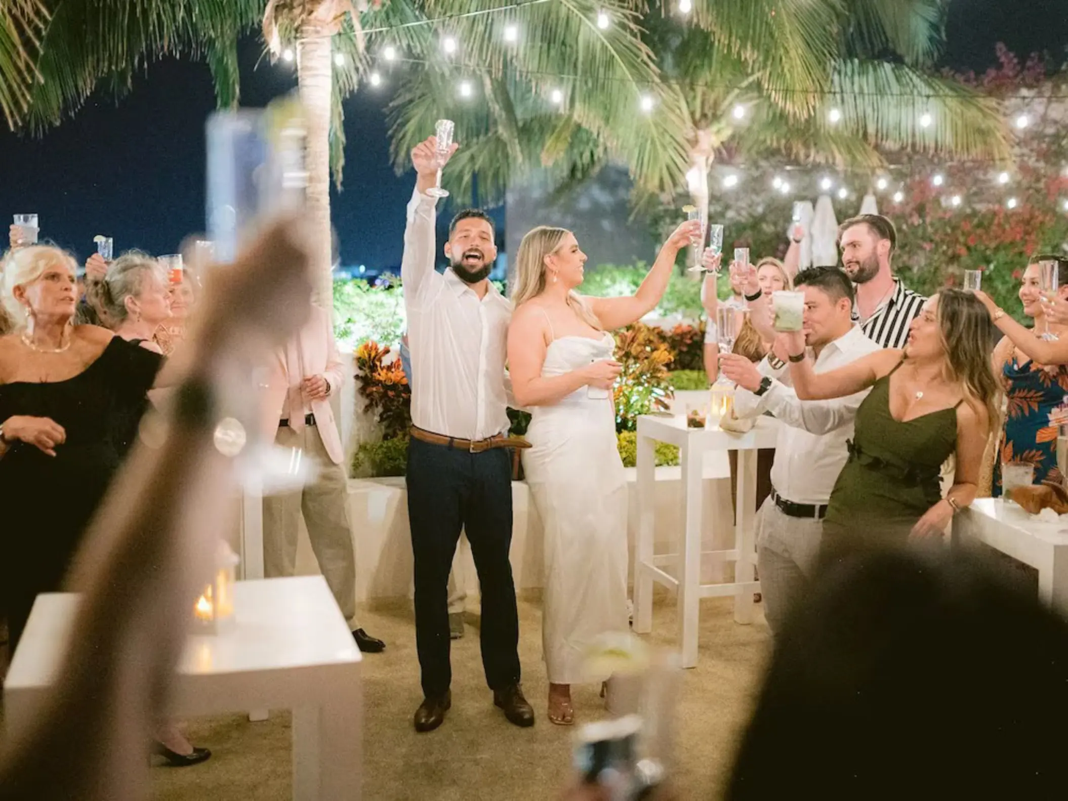 A joyful couple raising a toast at their outdoor wedding celebration at The Reef 28, surrounded by cheering guests at night.
