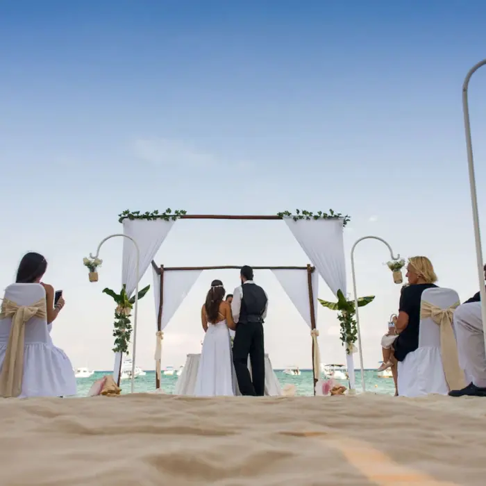 A couple getting married under a white canopy on the beach, surrounded by seated guests.