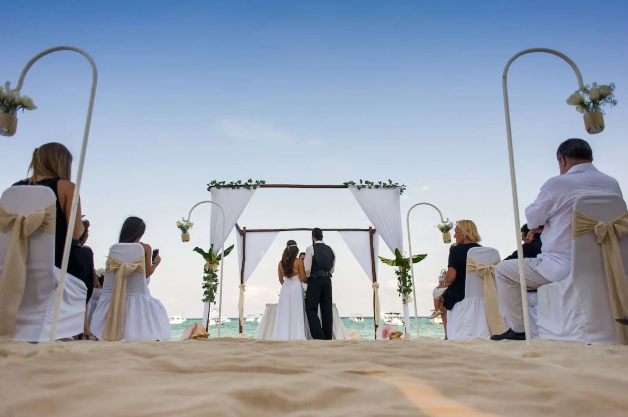 A couple getting married under a white canopy on the beach, surrounded by seated guests.