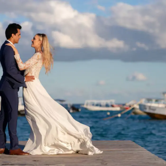 Bride and groom posing on a wooden pier with boats and the ocean in the background.