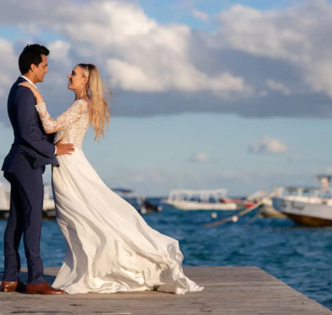 Bride and groom posing on a wooden pier with boats and the ocean in the background.
