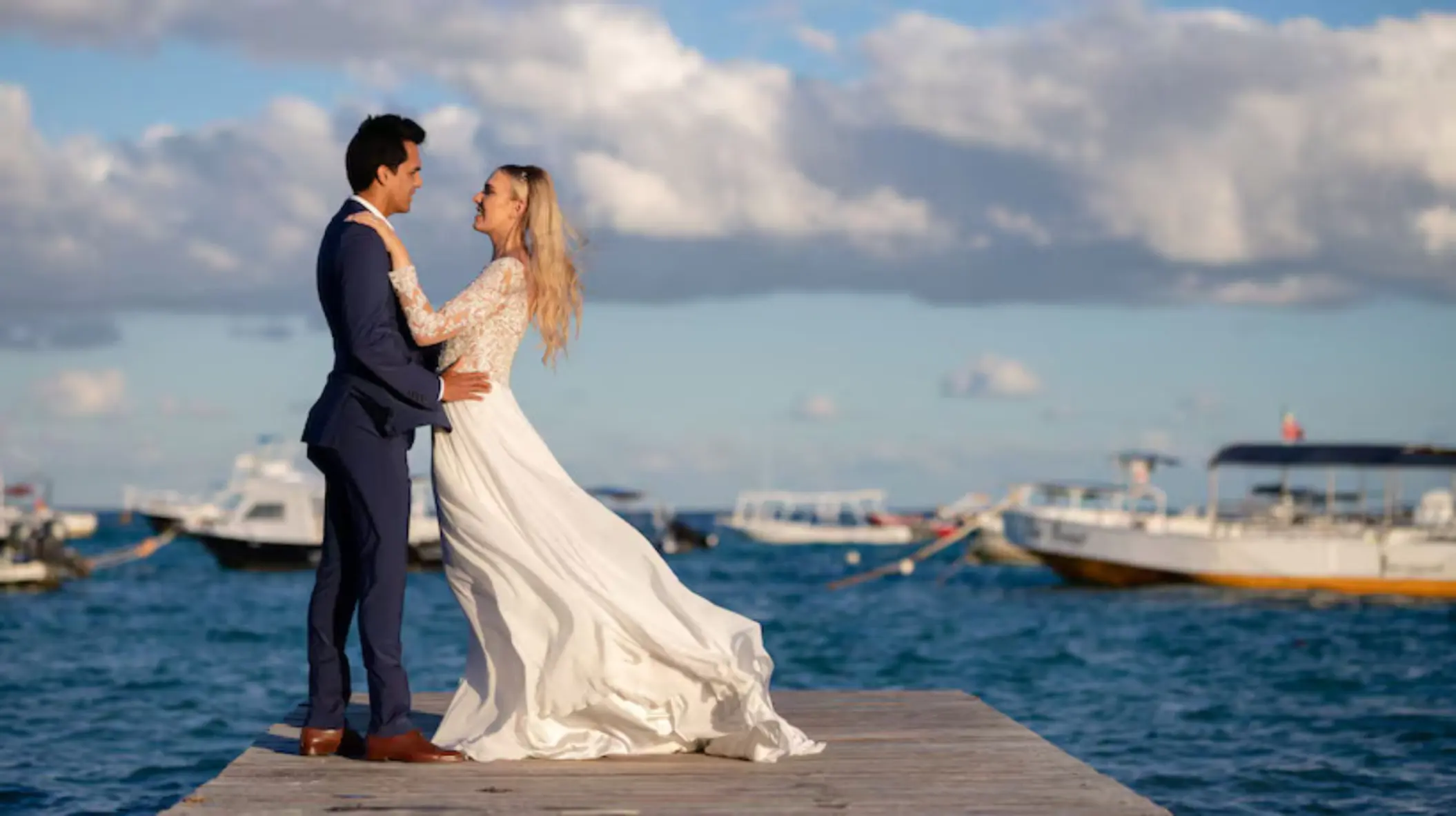 Bride and groom posing on a wooden pier with boats and the ocean in the background.