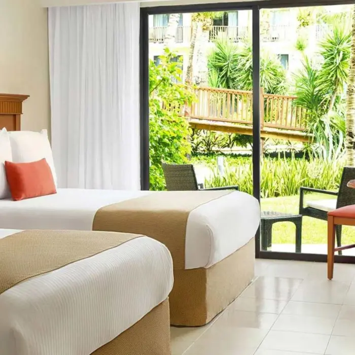 A bright hotel room with two beds and a sliding glass door opening to a garden view.