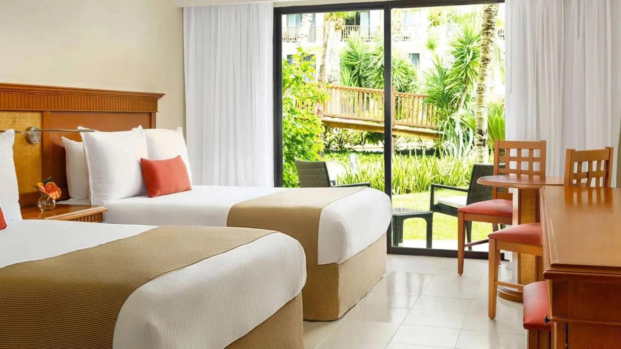 A bright hotel room with two beds and a sliding glass door opening to a garden view.