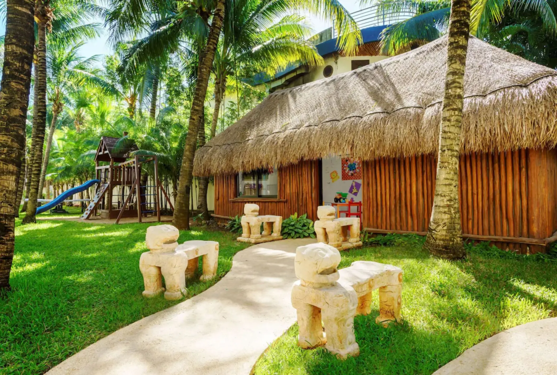 A children's playground with a slide and a small thatched-roof hut surrounded by palm trees.