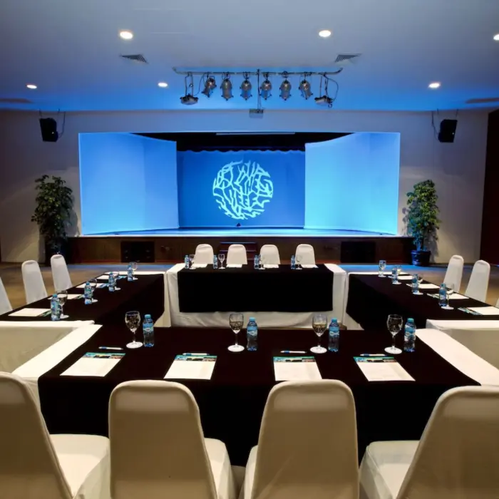 A U-shaped conference table with chairs facing a stage and projector screen.