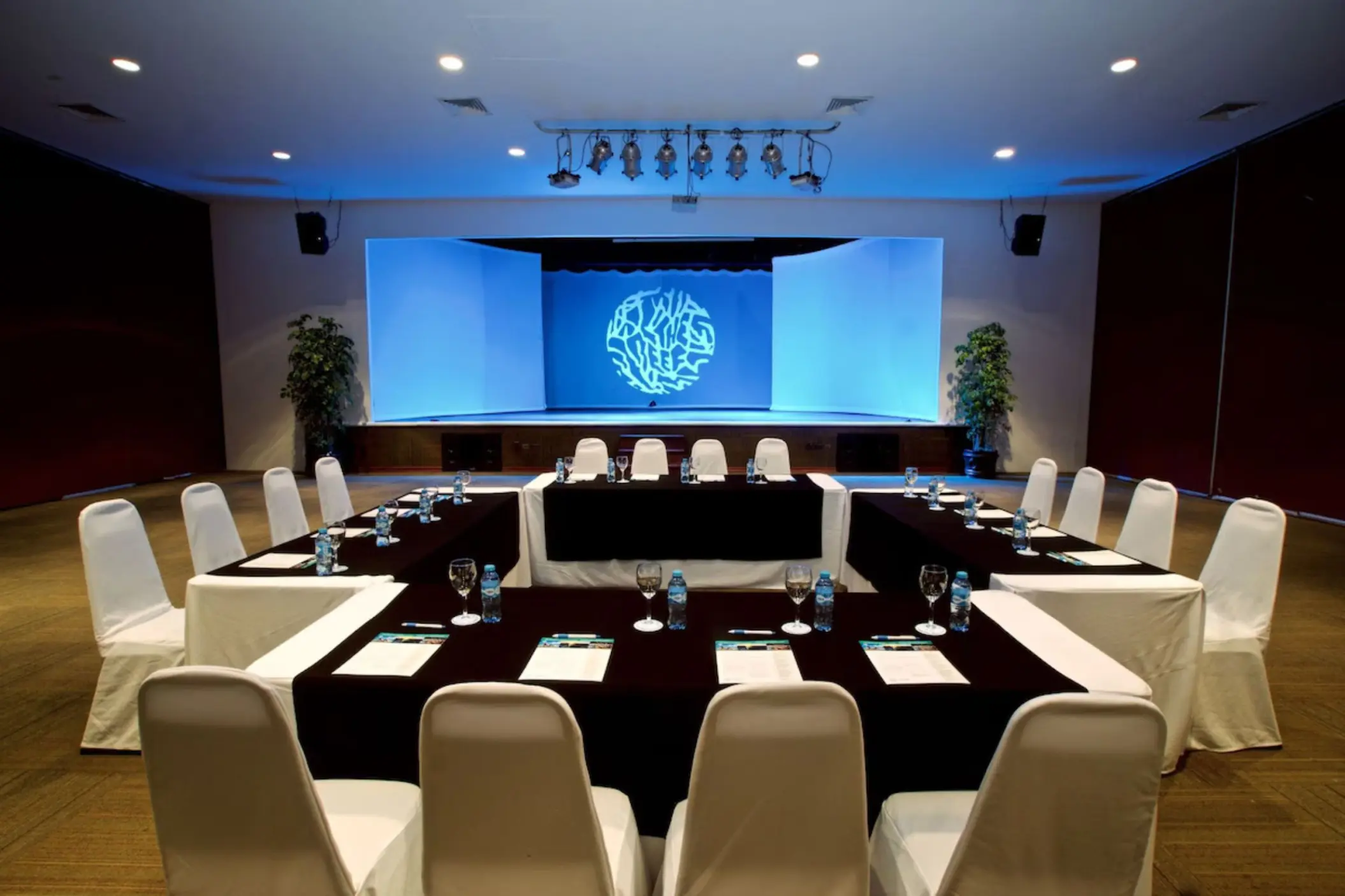 A U-shaped conference table with chairs facing a stage and projector screen.