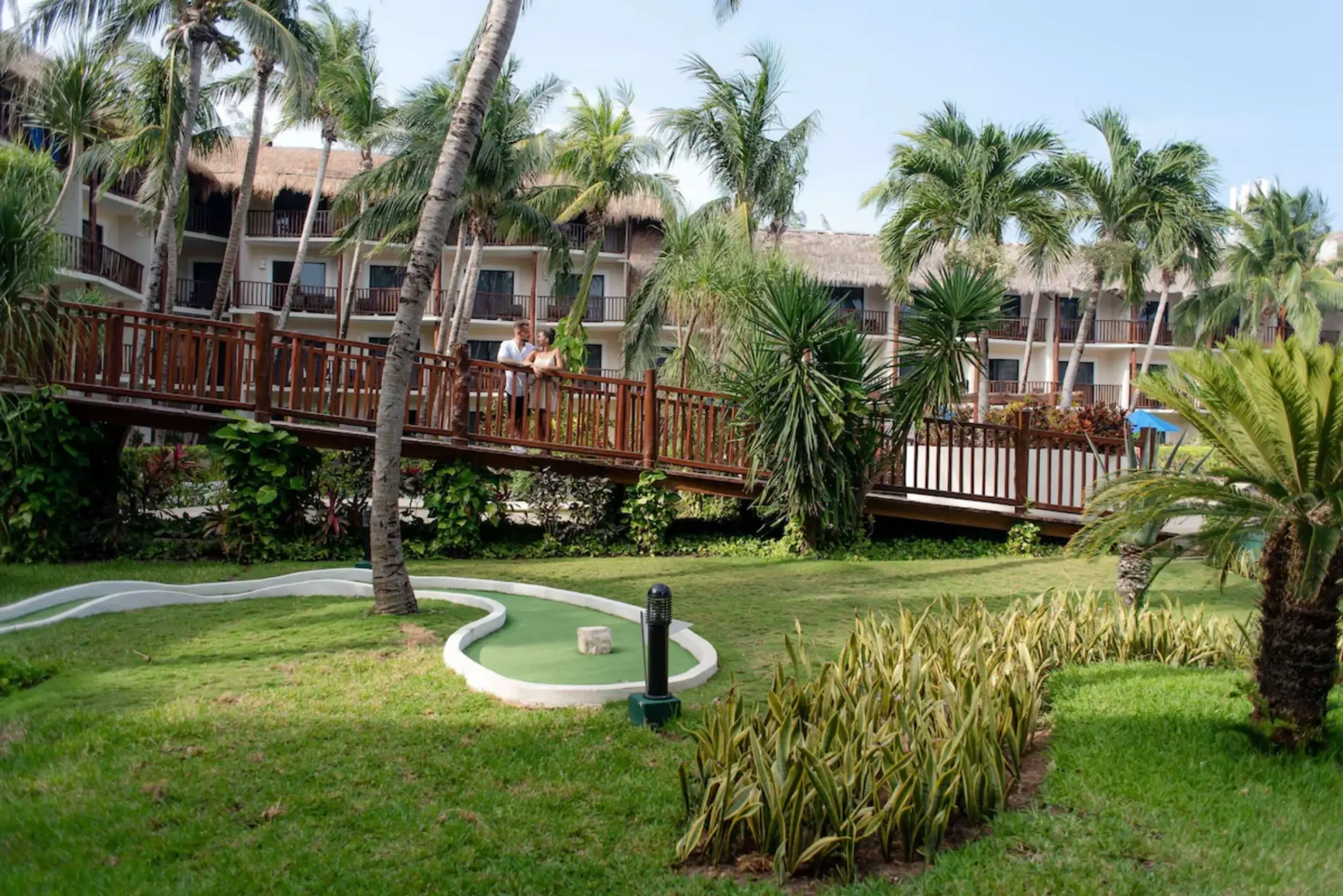 A mini-golf course surrounded by lush green lawn and palm trees with a couple in the background.