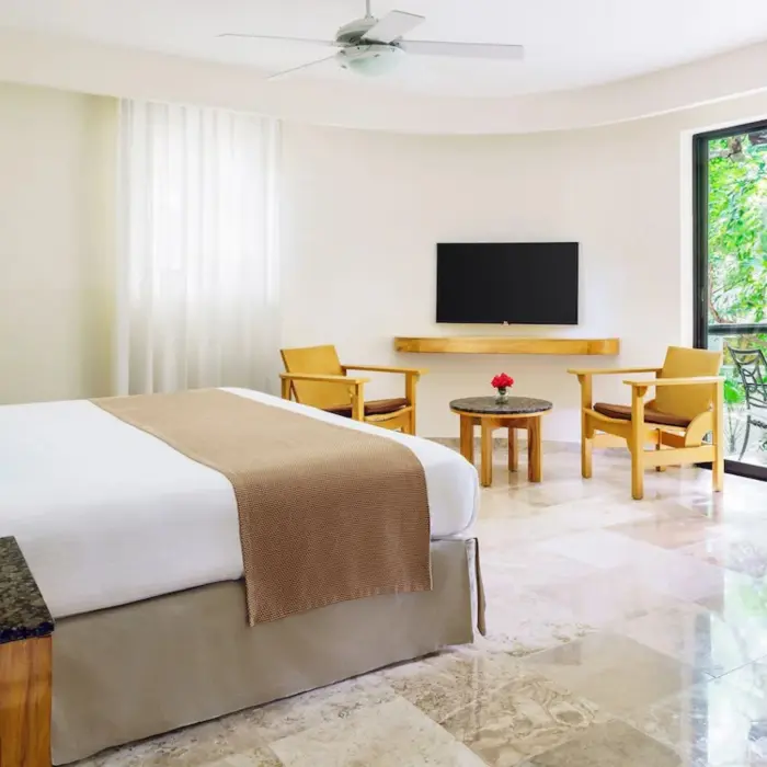 A bright hotel bedroom at The Reef Playacar with a large bed, seating area, and a lush garden view from the window.