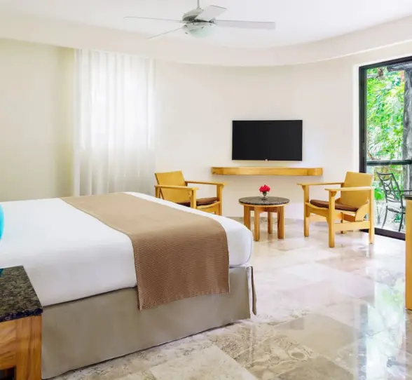 A bright hotel bedroom at The Reef Playacar with a large bed, seating area, and a lush garden view from the window.