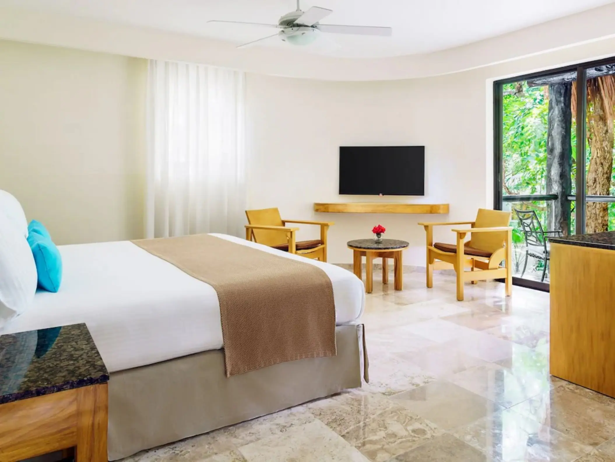 A bright hotel bedroom at The Reef Playacar with a large bed, seating area, and a lush garden view from the window.