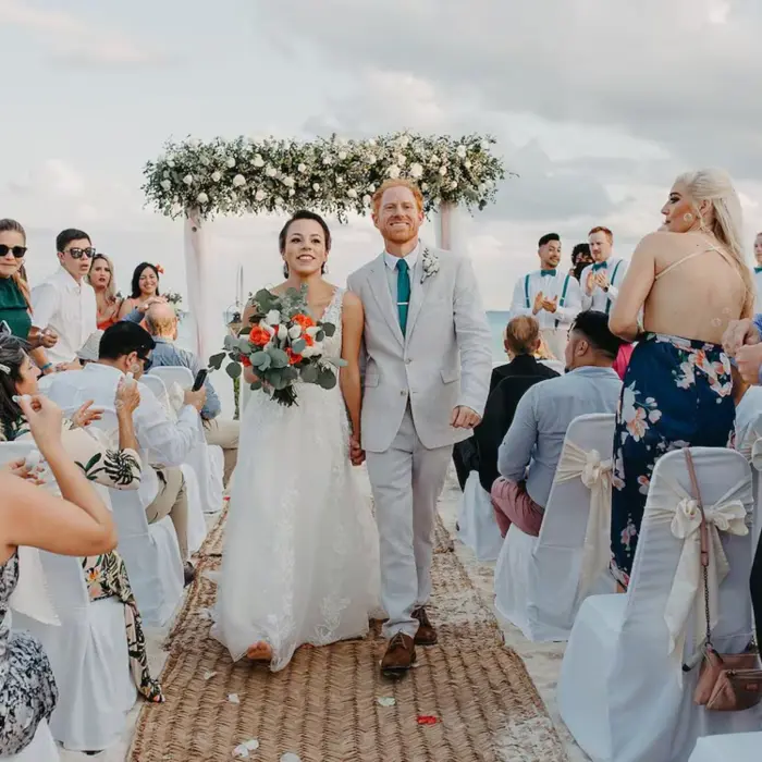 A joyful bride and groom walking down an aisle on the beach, surrounded by smiling guests at their wedding at The Reef Playacar.