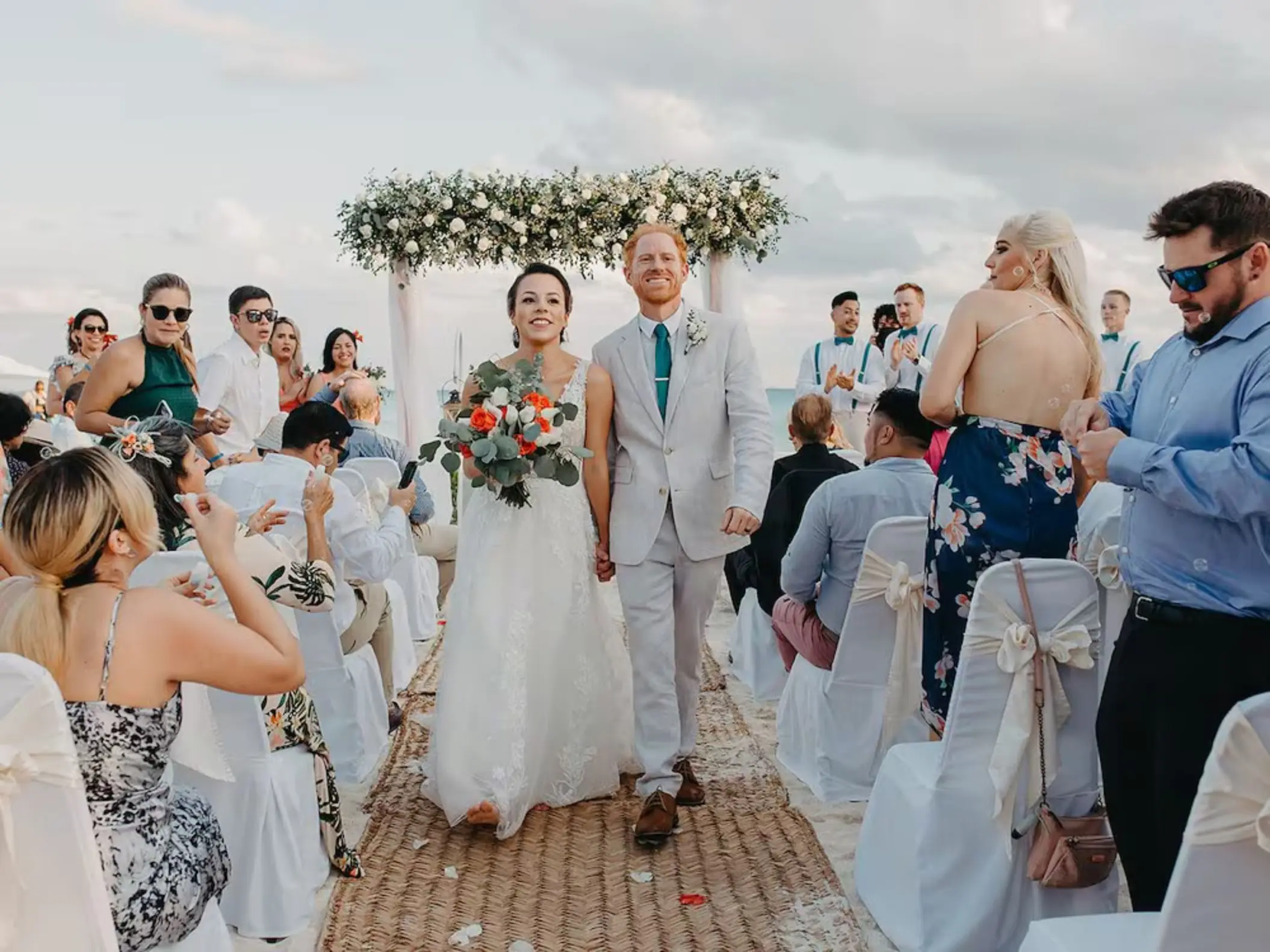A joyful bride and groom walking down an aisle on the beach, surrounded by smiling guests at their wedding at The Reef Playacar.
