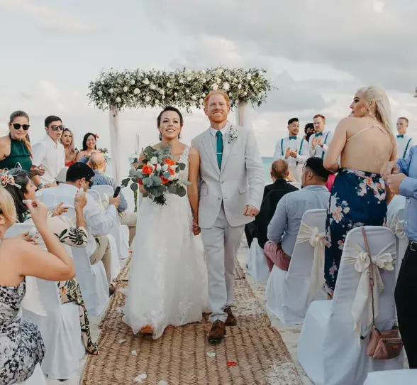 A joyful bride and groom walking down an aisle on the beach, surrounded by smiling guests at their wedding at The Reef Playacar.