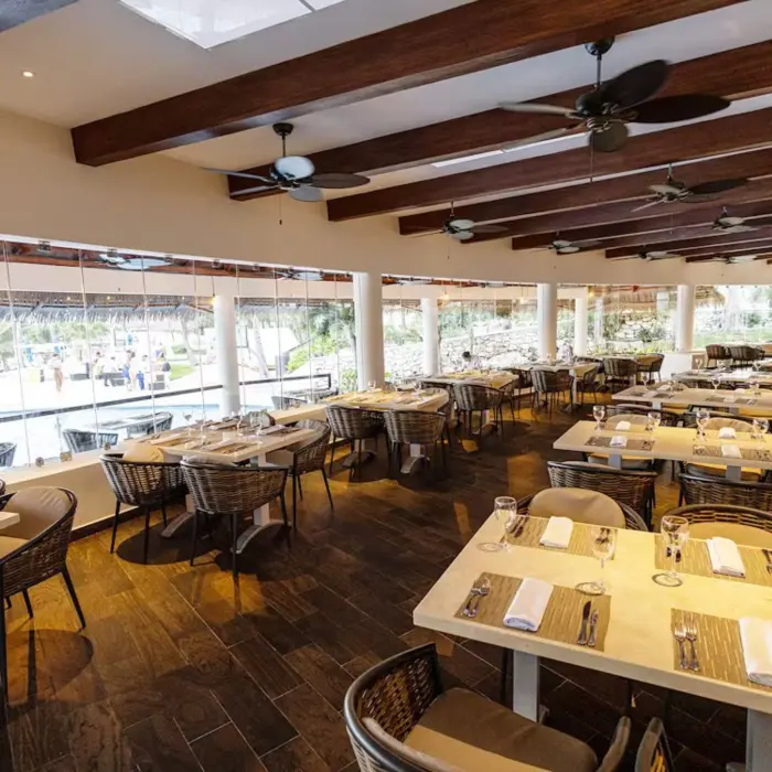 Interior of the spacious Chulavista restaurant at The Reef Playacar, featuring many dining tables and large windows overlooking a pool area.