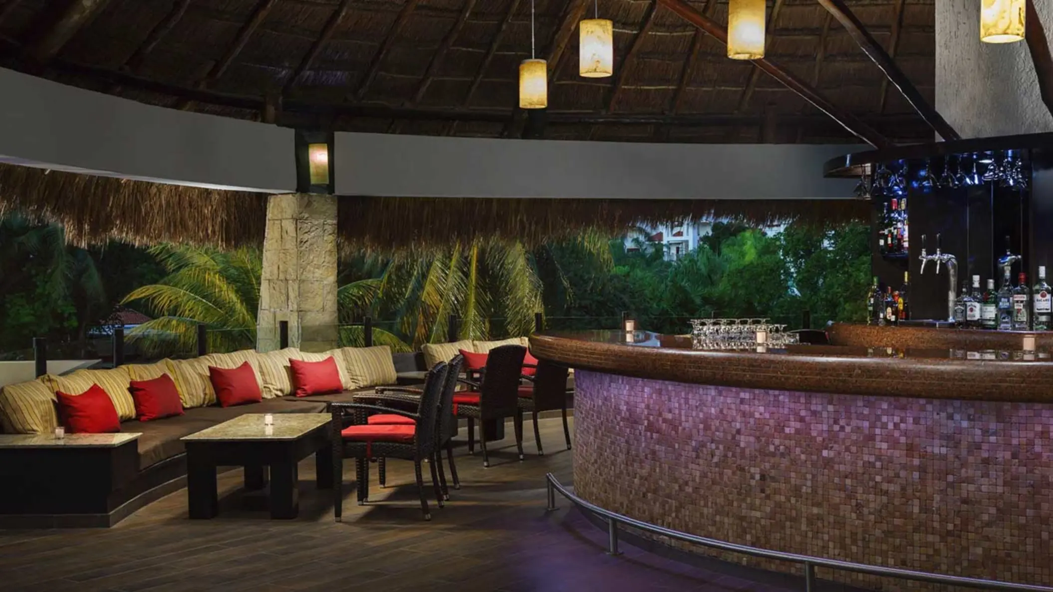 inviting interior of Papos Bar at The Reef Playacar, featuring a well-stocked bar, cozy seating with red cushions, and a thatched roof.